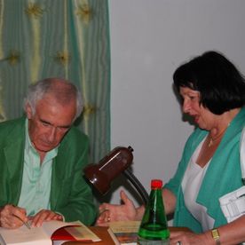Älterer Herr unterzeichnet Buch, Dame steht daneben